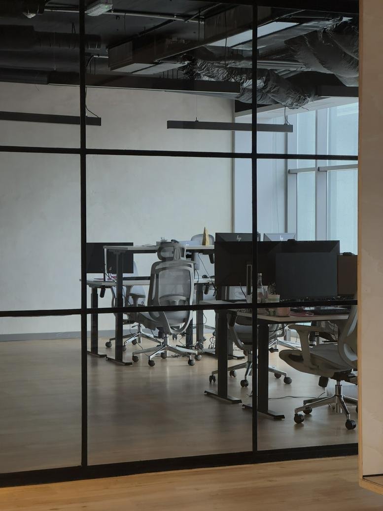 View through a black-framed glass partition into a modern workspace with dual-monitor desks.
