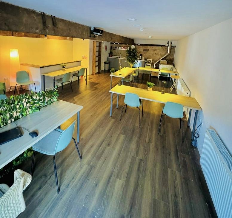 Wide-angle view of a shared office space with multiple desks, blue chairs, and exposed wooden ceiling beams.